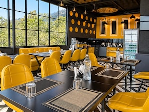 A restaurant with yellow chairs and tables, a bar area, and a view of greenery through large windows. - Hotel Celestial Ganges (Rishikesh)