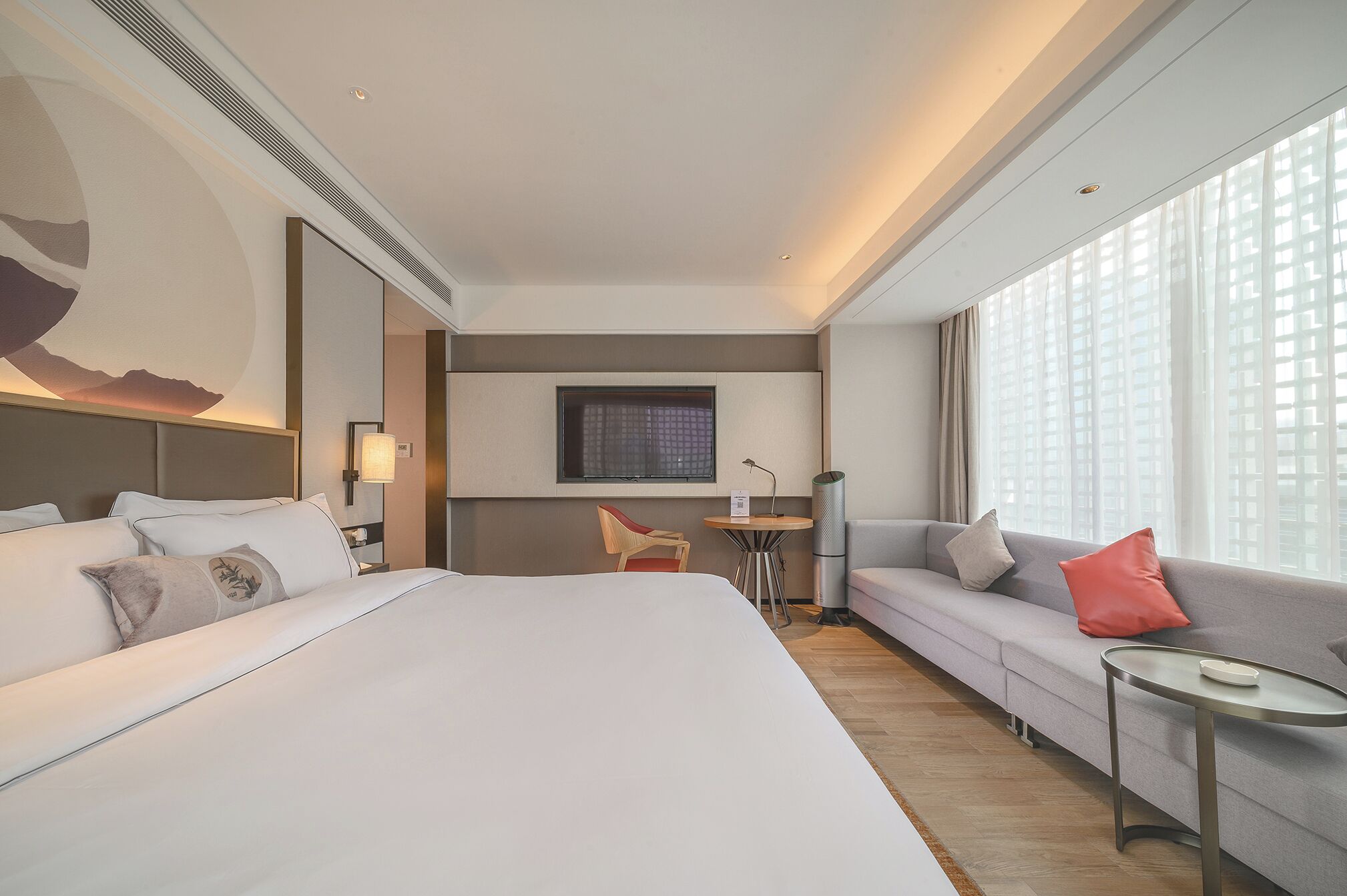 A hotel room with a large bed, a sofa, a small table, a chair, a TV, and a window with blinds.