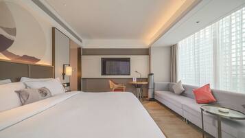 Habitación de hotel con una cama grande, un sofá, una mesita, una silla, un televisor y una ventana con persianas.