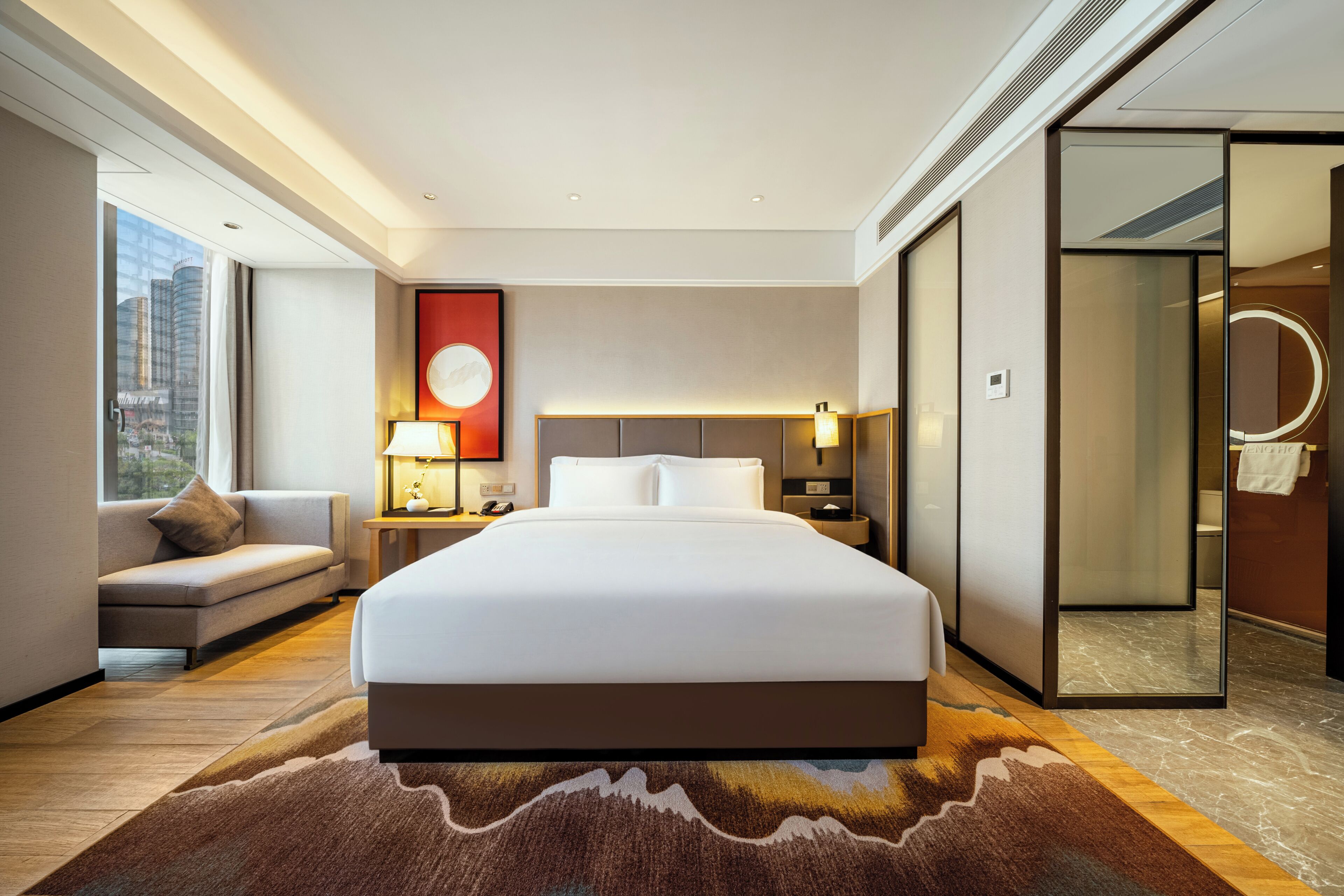 Ett modernt hotellrum med en stor säng, en soffa, ett sängbord, en lampa och utsikt över staden genom ett stort fönster.