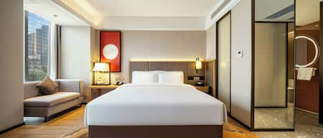 A modern hotel room with a large bed, a sofa, a nightstand, a lamp, and a view of the city through a large window.