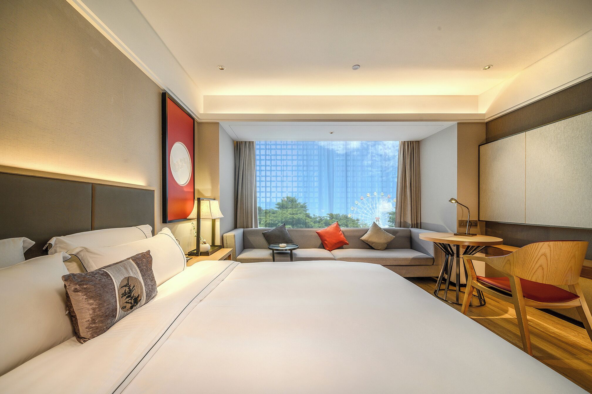 A hotel room with a large bed, a sofa, a small table, and a chair. There is a window with a view of buildings and trees.