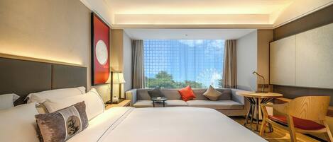 A hotel room with a large bed, a sofa, a small table, and a chair. There is a window with a view of buildings and trees.