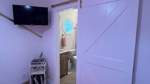 Ein Schlafzimmer mit einem rosa, flauschigen Teppich, einem an der Wand montierten Fernseher, einer Schiebetür zu einem Badezimmer und einer kleinen Ablage mit Gegenständen.
