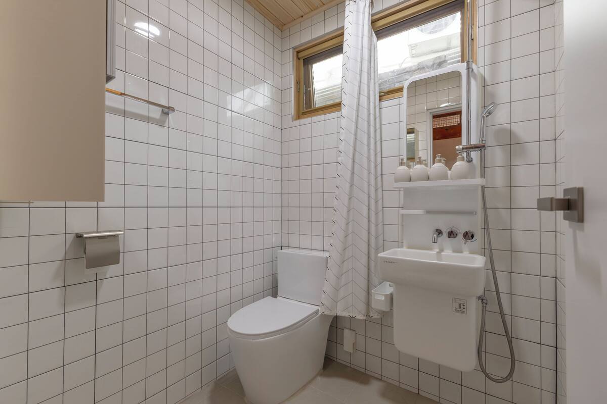 A bathroom with white tiled walls, a toilet, a shower with a handheld showerhead, and a mirror above the sink.