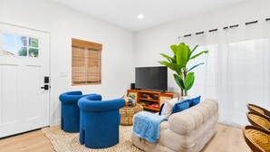 Un salon avec un canapé beige, des fauteuils bleus, un téléviseur et une grande plante.