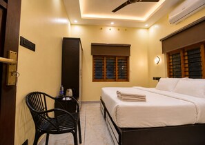 A hotel room with a bed, a desk, a chair, a window with blinds, and an air conditioning unit. - De Waves (Varkala)