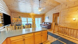 A wooden cabin interior with a kitchen area featuring a stone fireplace, wooden cabinets, and a large TV. - Lost on Purpose : Summer Memories Made Here | Cabin for 12 (Wetmore)