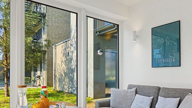 A modern living room with a grey sofa, a dining table set for four, and a large window offering a view of a brick building and greenery. - Seemomente 615 (Seenähe) by Interhome (Lembruch)