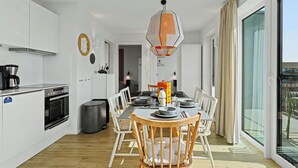 Moderni keittiö, jossa on valkoiset kaapit, ruokailutila puupöydällä ja -tuoleilla sekä suuri ikkuna, jossa on verhot.