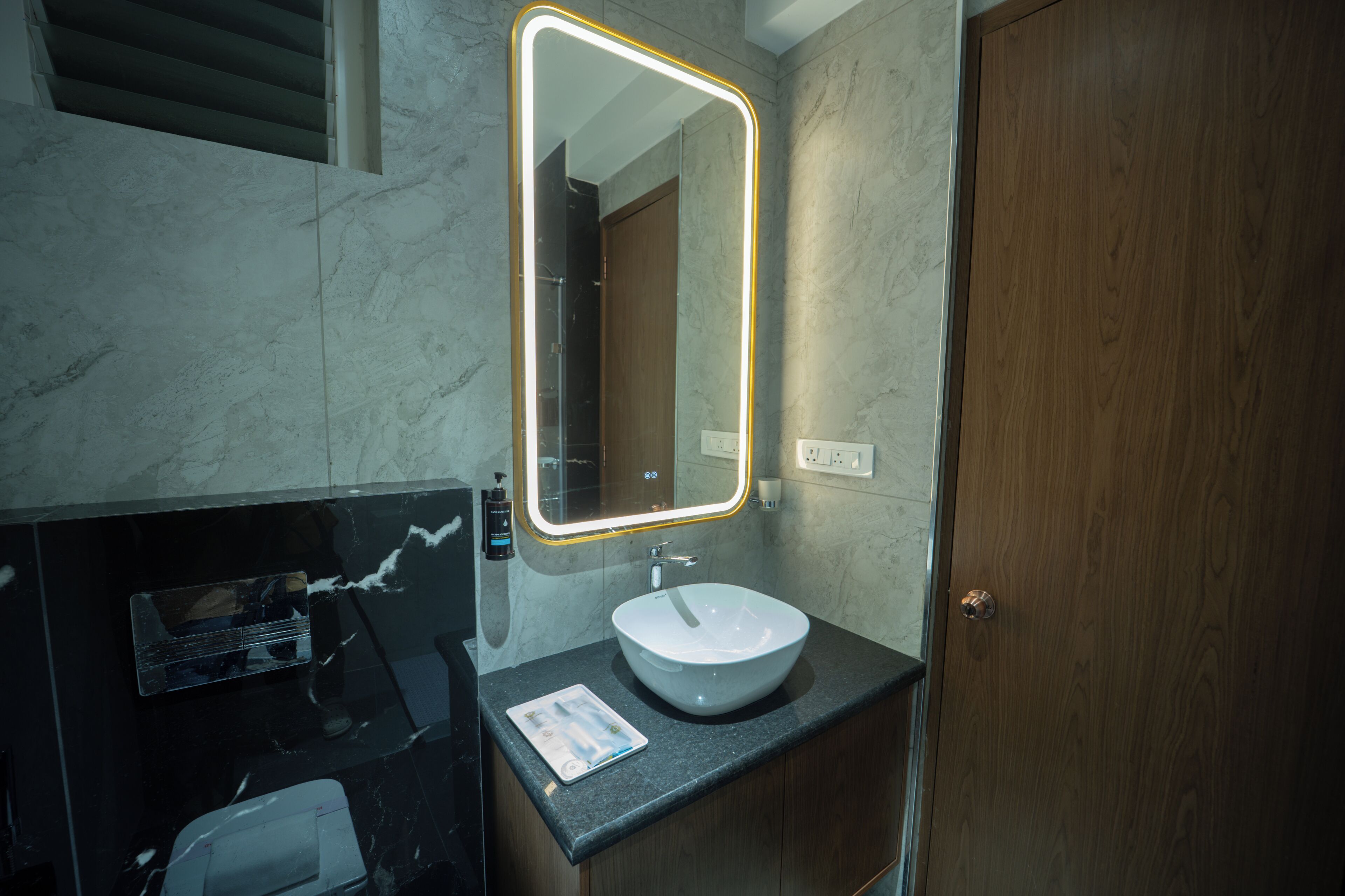 Un baño moderno con un lavabo de mármol, un espejo con marco iluminado y una puerta de madera.