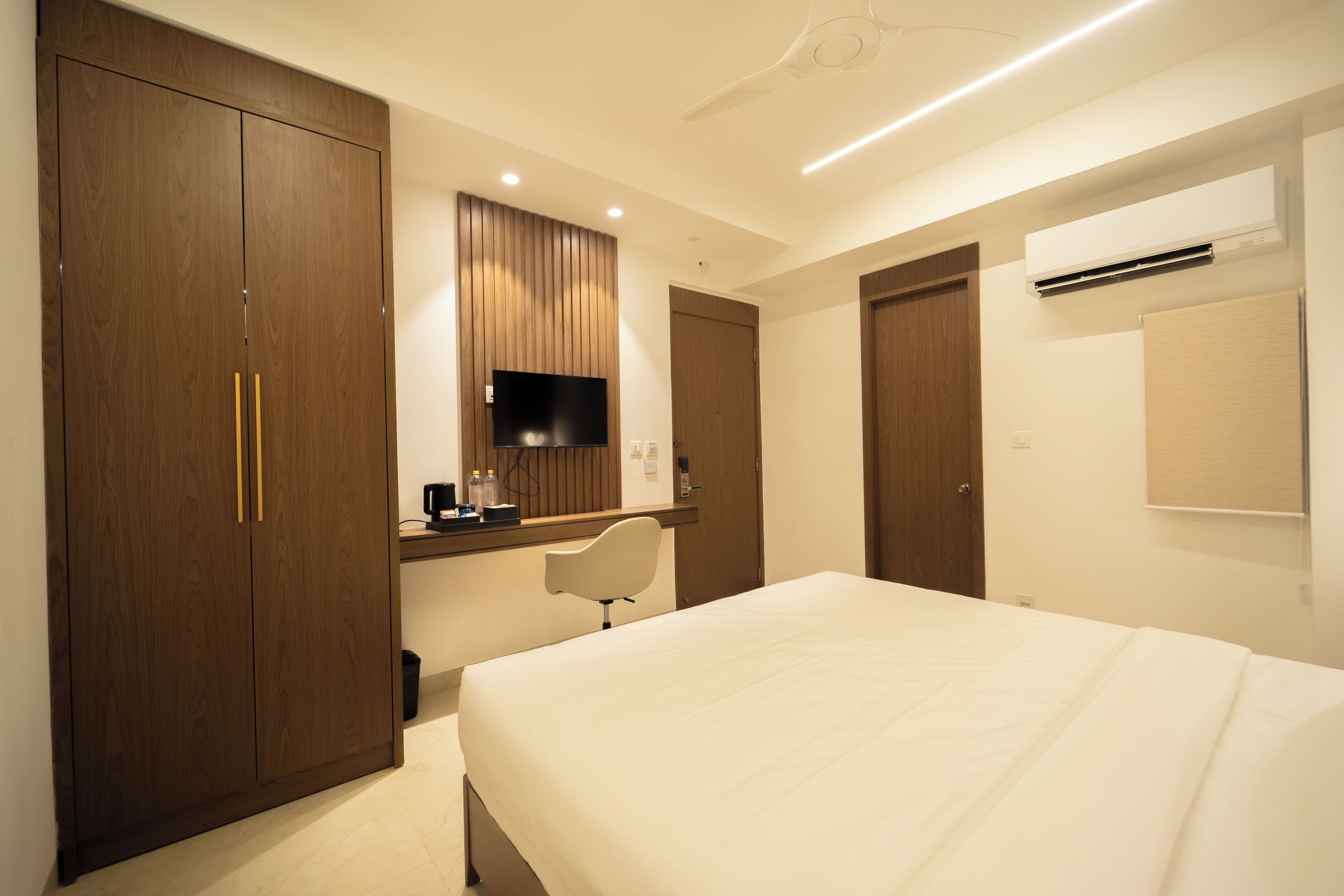 A hotel room with a large bed, a desk with a chair, a wardrobe, a television, and an air conditioning unit.
