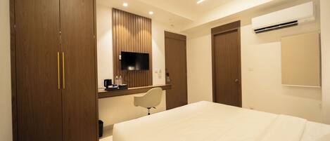 A hotel room with a large bed, a desk with a chair, a wardrobe, a television, and an air conditioning unit.