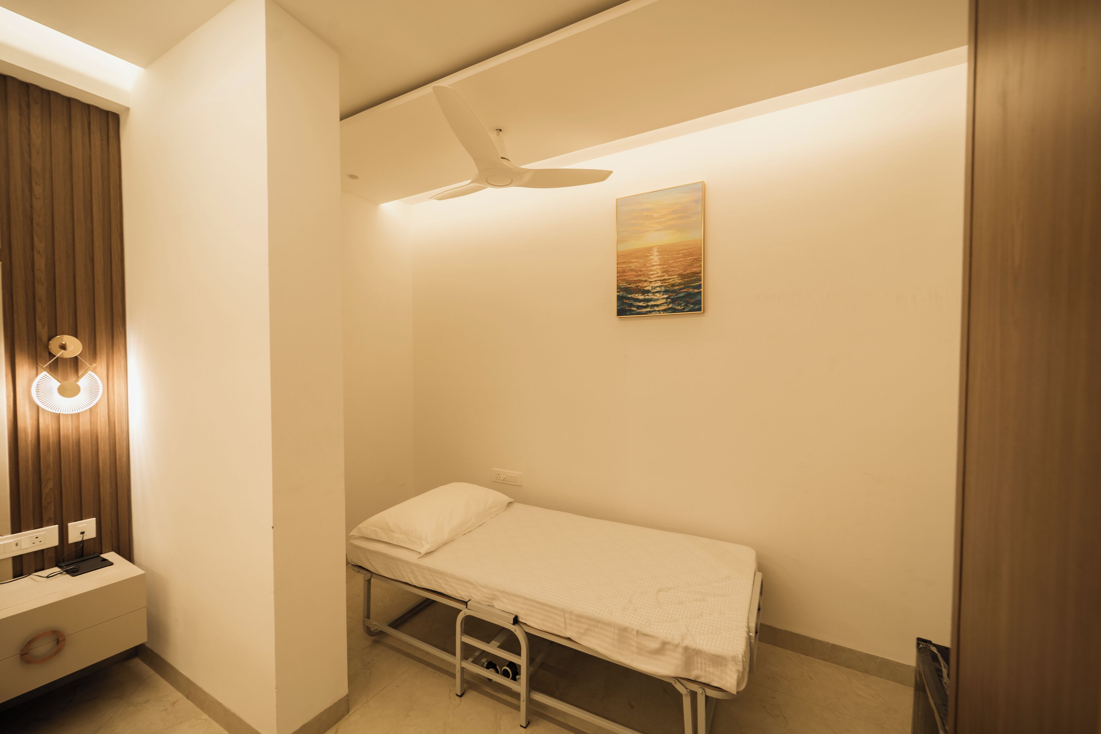 Habitación individual con cama, ventilador de techo, mesita de noche con lámpara, un cuadro en la pared y un armario de madera.