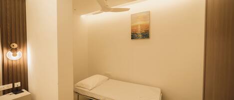 Habitación individual con cama, ventilador de techo, mesita de noche con lámpara, un cuadro en la pared y un armario de madera.