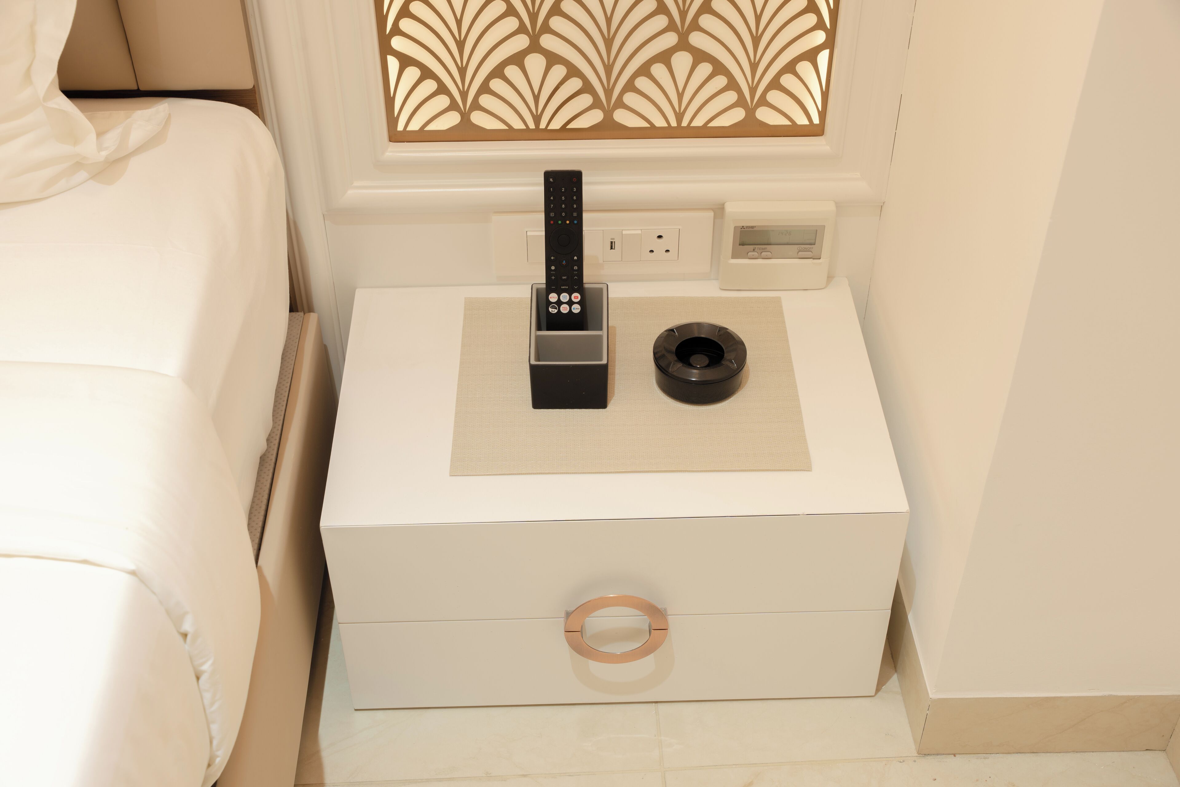 A neatly made bed with a white headboard, a bedside table with a remote control, a small black object, and a wall-mounted light switch.