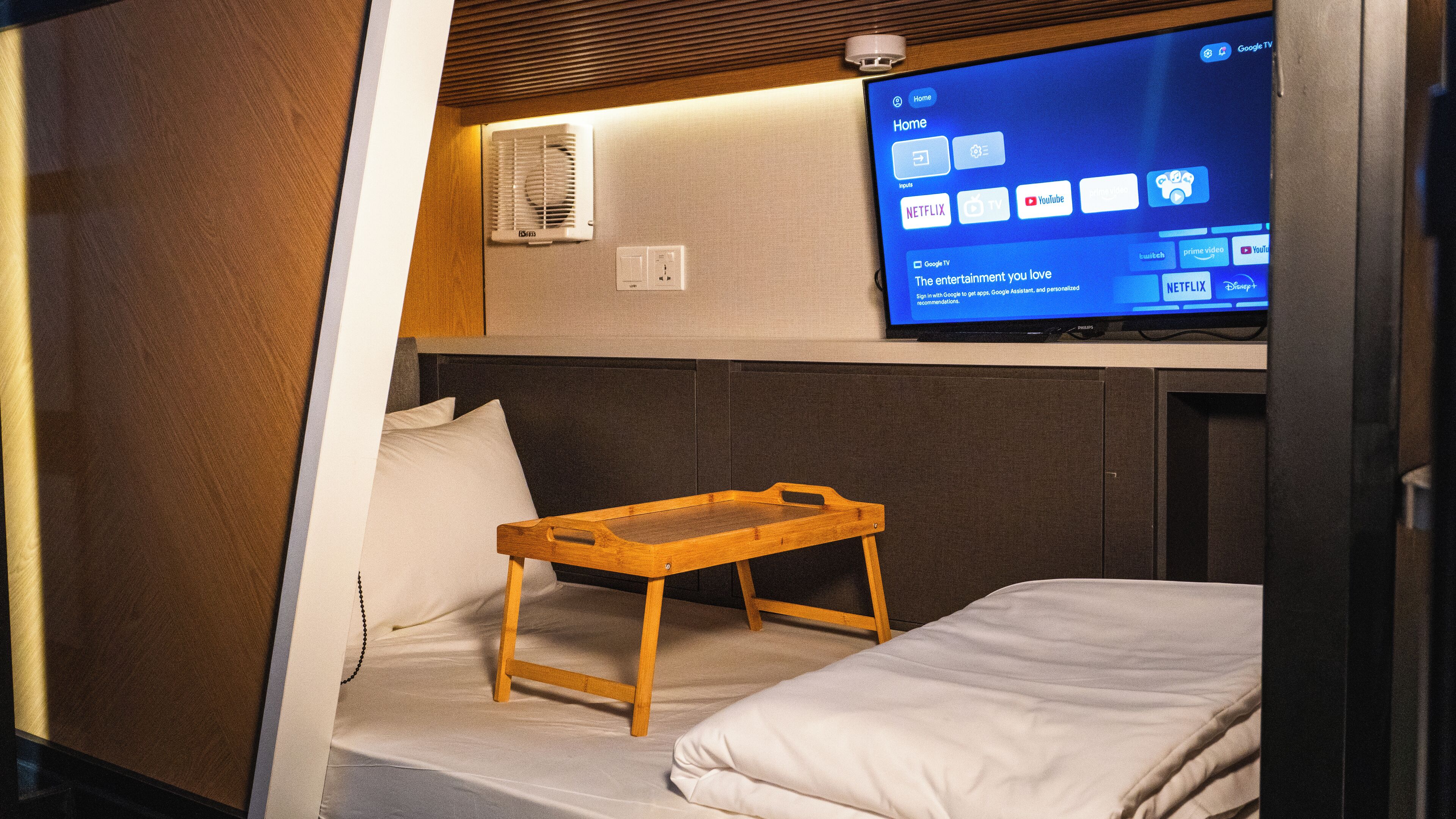 A compact room with a bed, a small wooden table, and a television displaying a home screen.