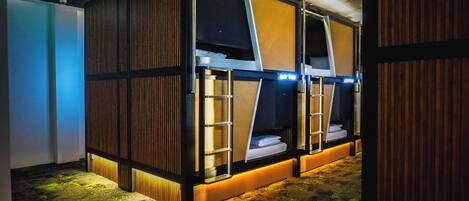A room with bunk beds, each with a ladder and a small shelf. The room has a wooden floor and a ceiling with recessed lighting.