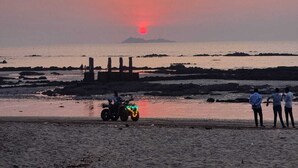 Escena de playa con un atardecer, un buggy de dunas y tres personas de pie en la arena.