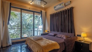 Habitación de hotel con un ventanal grande, cama con cabecera, mesita de noche con lámpara y aire acondicionado instalado en la pared.