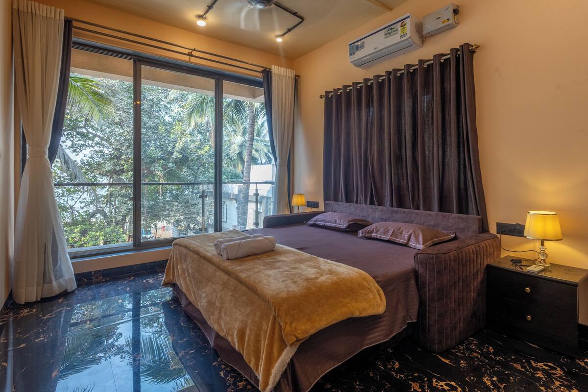 A hotel room with a large window, a bed with a headboard, a bedside table with a lamp, and a wall-mounted air conditioner.