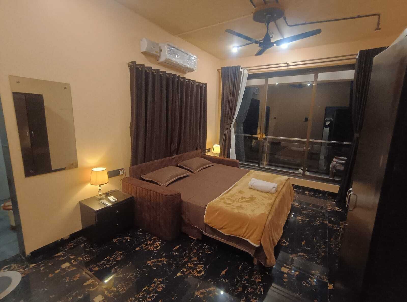Habitació d'hotel amb un llit, una tauleta de nit, una làmpada, un ventilador de sostre i una finestra amb cortines.