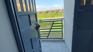 Ein Balkon mit einem Metallgitter, Fliesenboden und Blick auf ein grünes Feld sowie Gebäude in der Ferne.