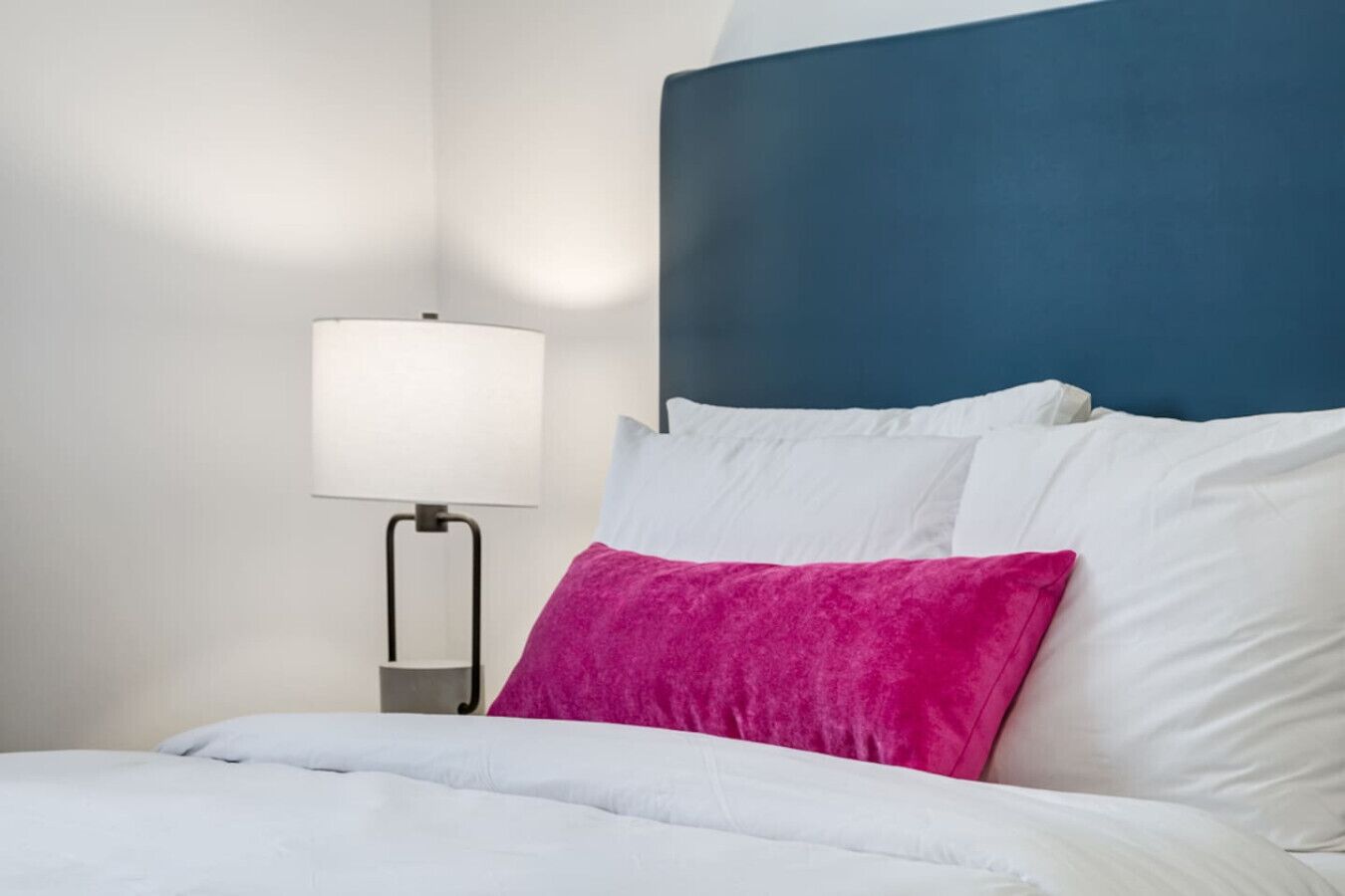 Una cama bien hecha con ropa blanca y una única almohada rosa brillante.