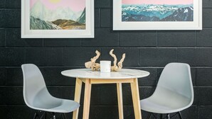 Une pièce avec un mur en briques noires, deux chaises grises, une petite table ronde sur laquelle se trouve une tasse blanche, et deux tableaux encadrés représentant des montagnes et un coucher de soleil.