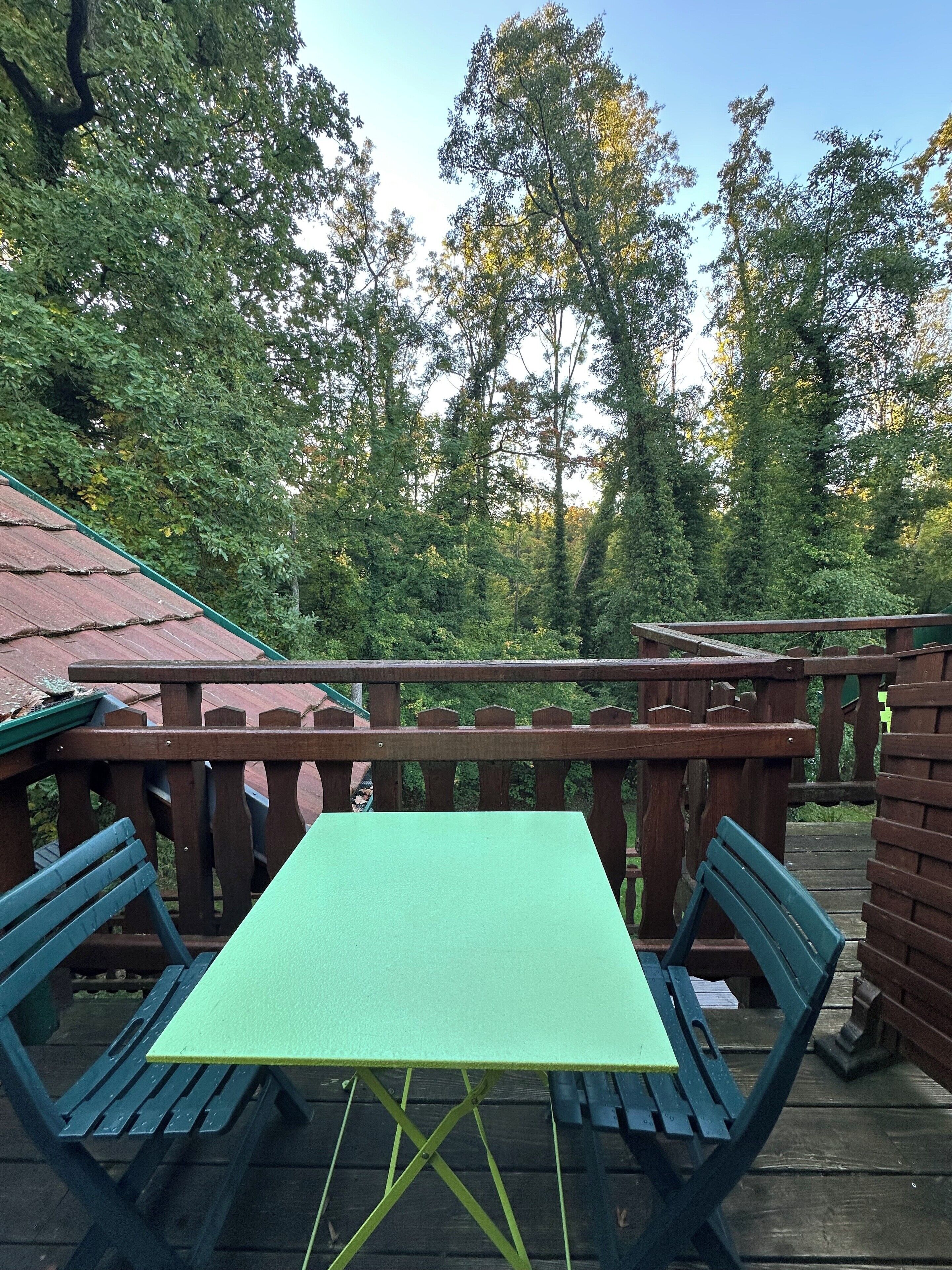 Uma mesa verde e duas cadeiras azuis num deck de madeira, com vista para árvores e um céu limpo.