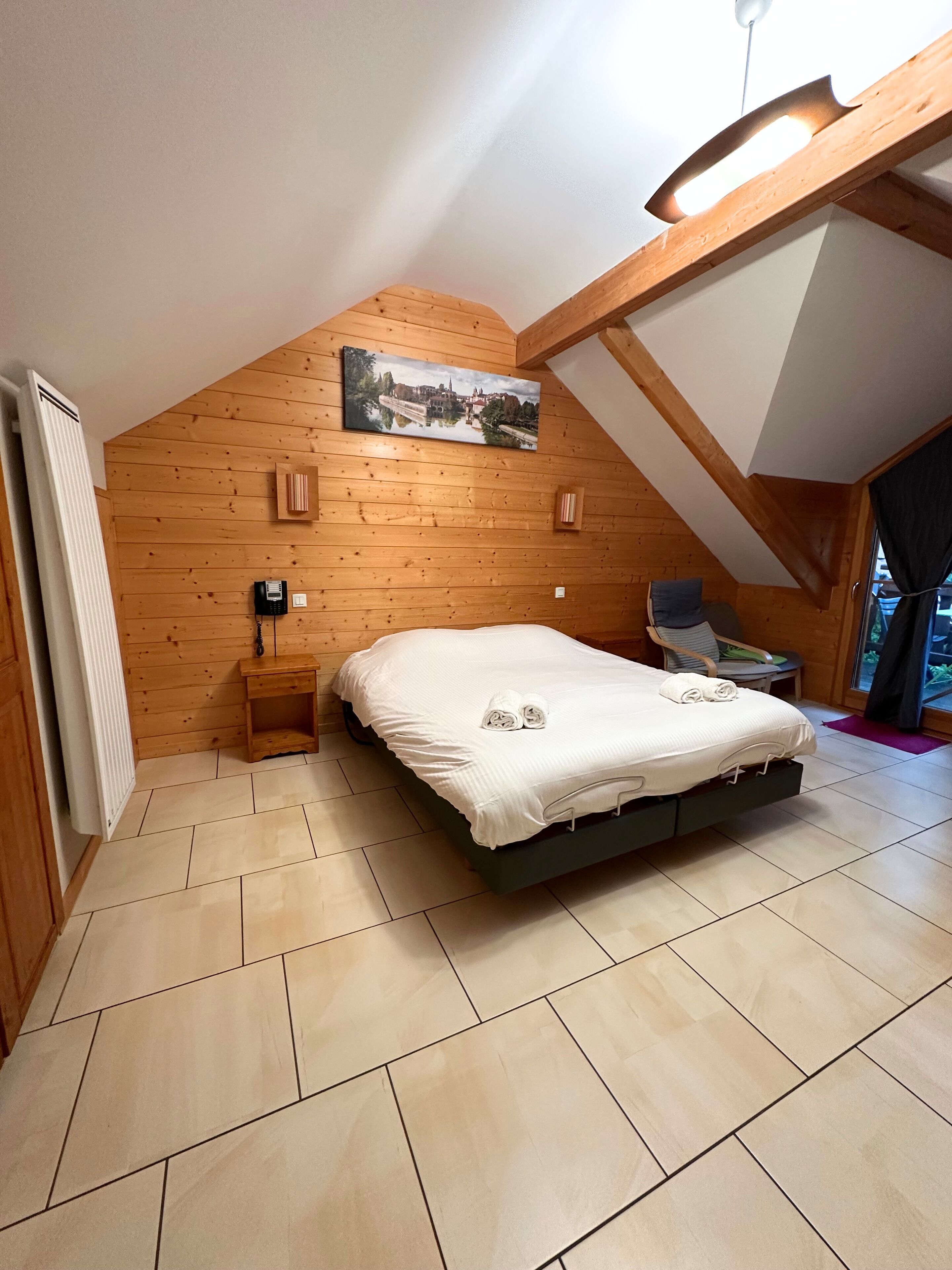 Ein Schlafzimmer mit schrägen Wänden, Holzwandverkleidung, einem Bett mit weißer Bettwäsche, einem Nachttisch, einem Sessel und einem Fenster mit Vorhängen.