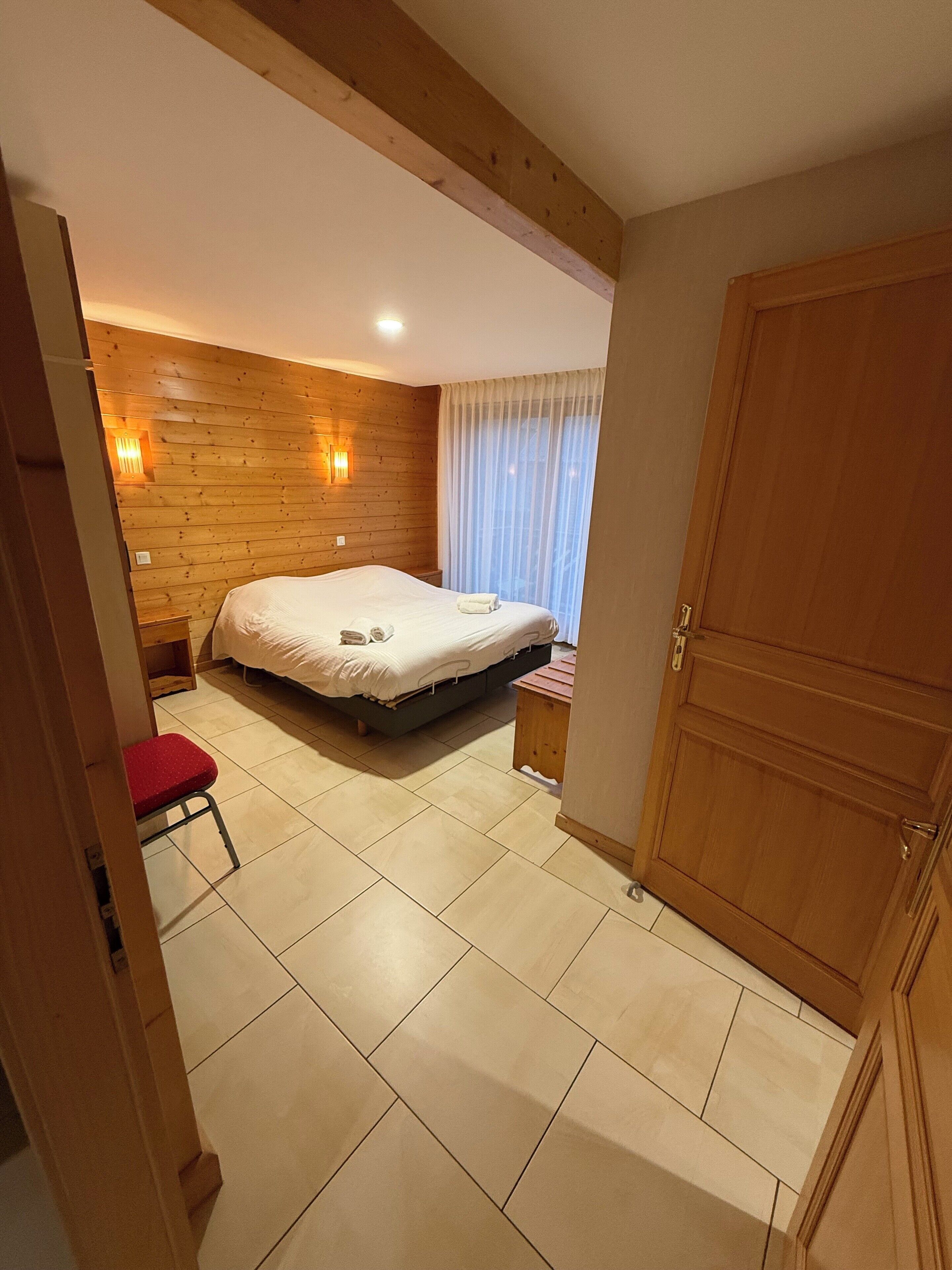 Un dormitorio con una cama, una pared de madera, un taburete pequeño y una puerta.