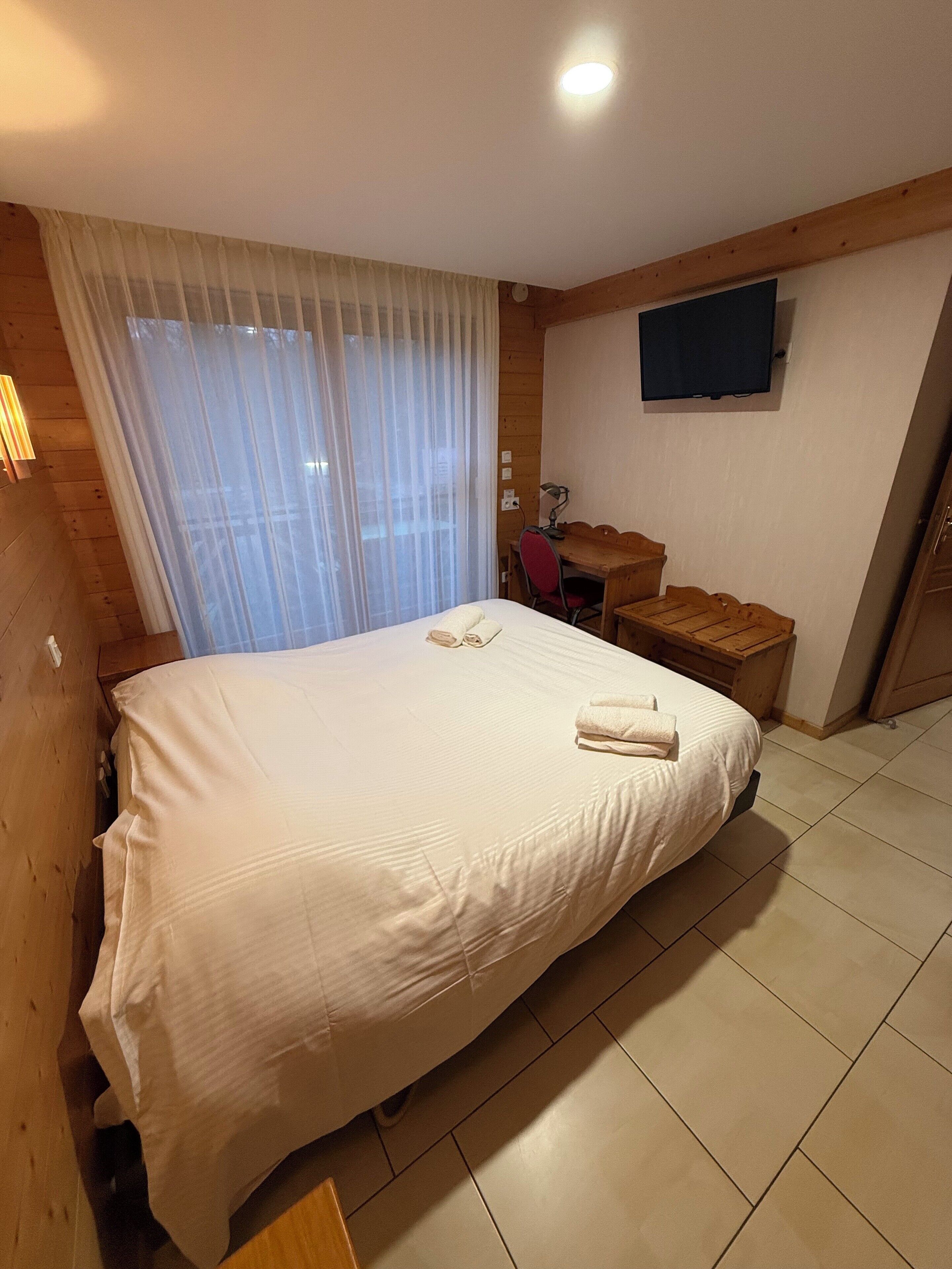 A hotel room with a large bed, a desk with a chair, a TV mounted on the wall, and a window with sheer curtains.