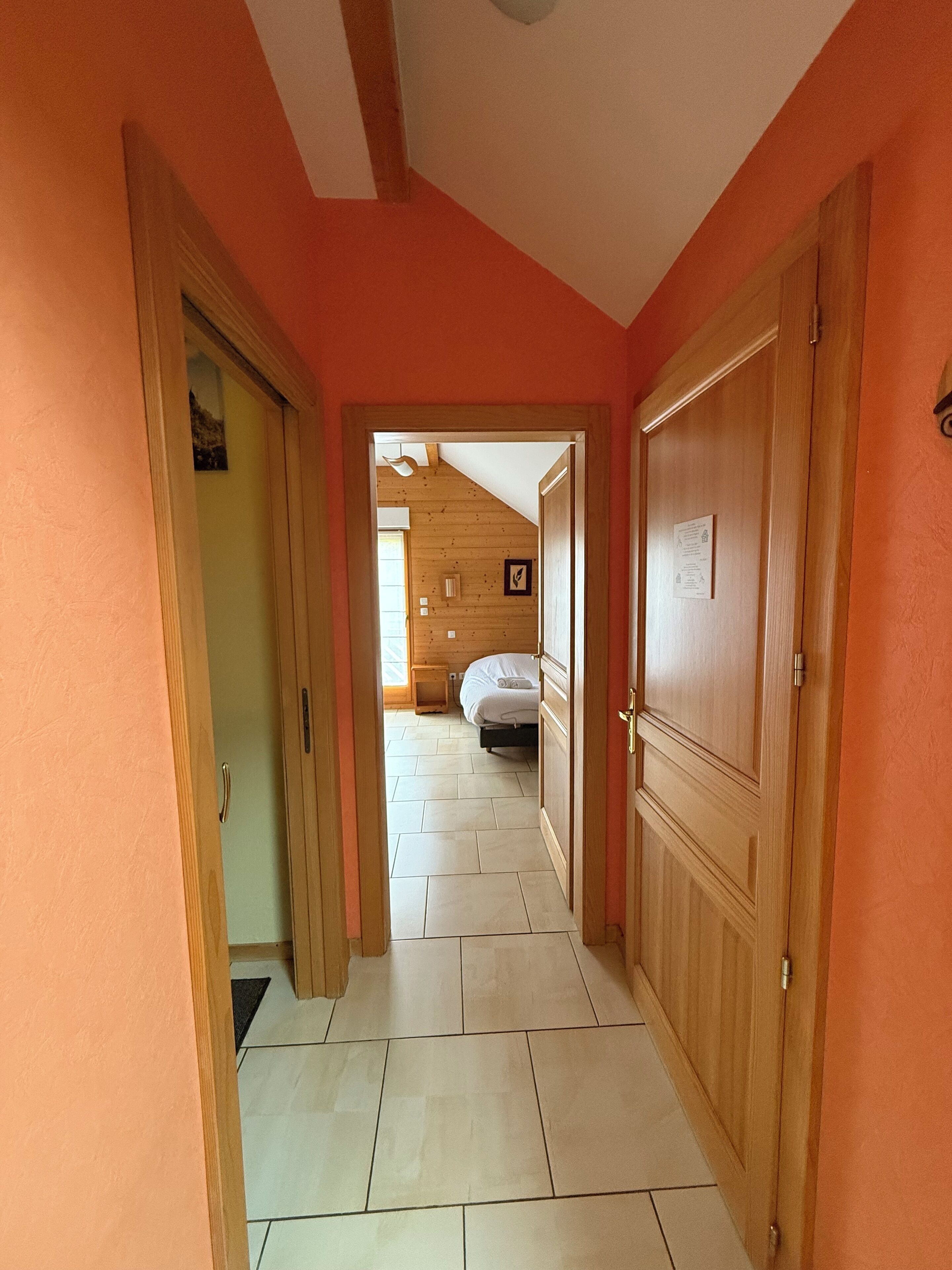 A hallway with wooden doors and a tiled floor leading to a bedroom with a bed and a nightstand.