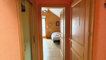 A hallway with wooden doors and a tiled floor leading to a bedroom with a bed and a nightstand.