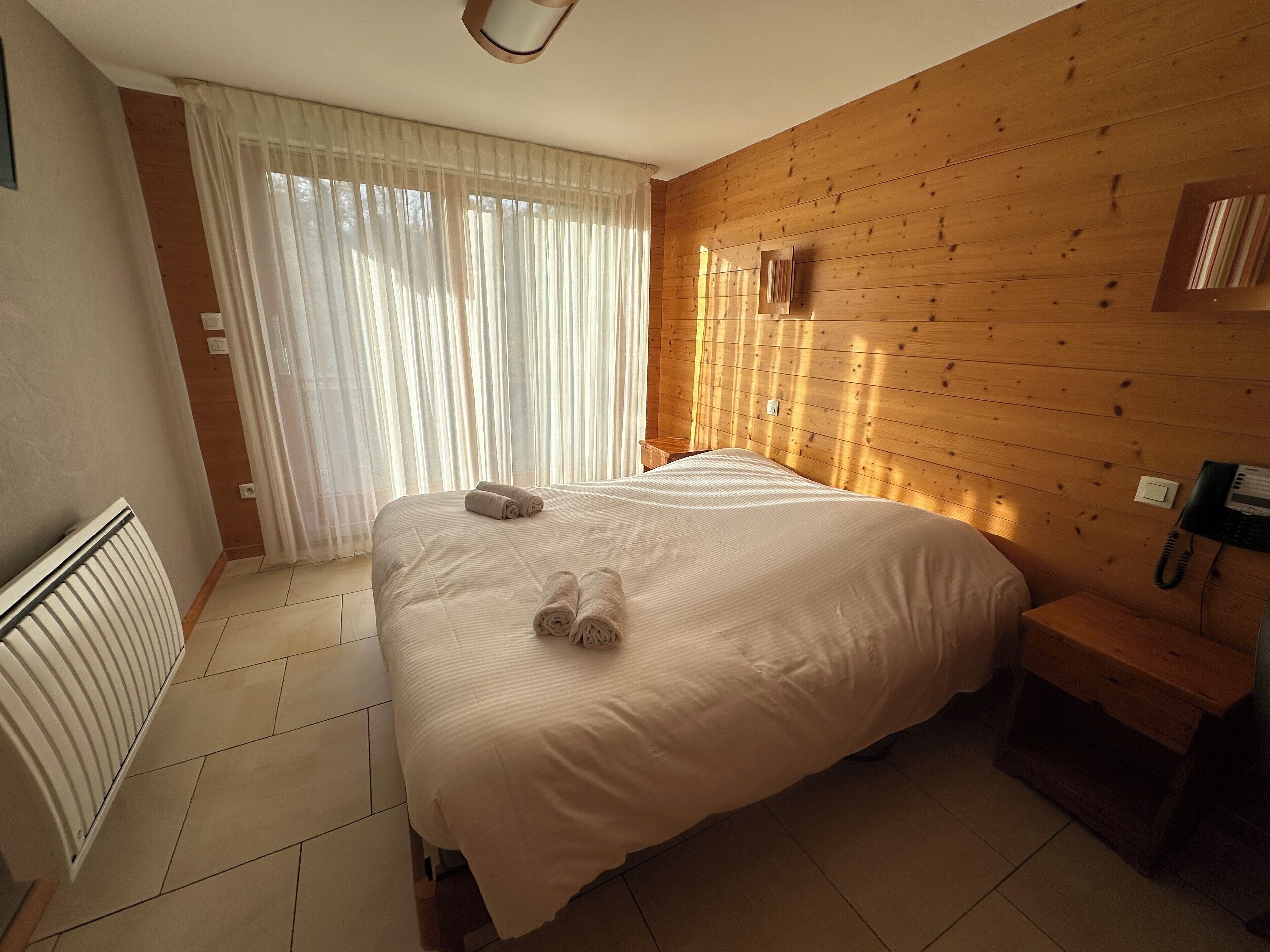 Quarto de hotel com uma cama grande, cabeceira de madeira, telefone ao lado da cama e uma janela com cortinas finas.