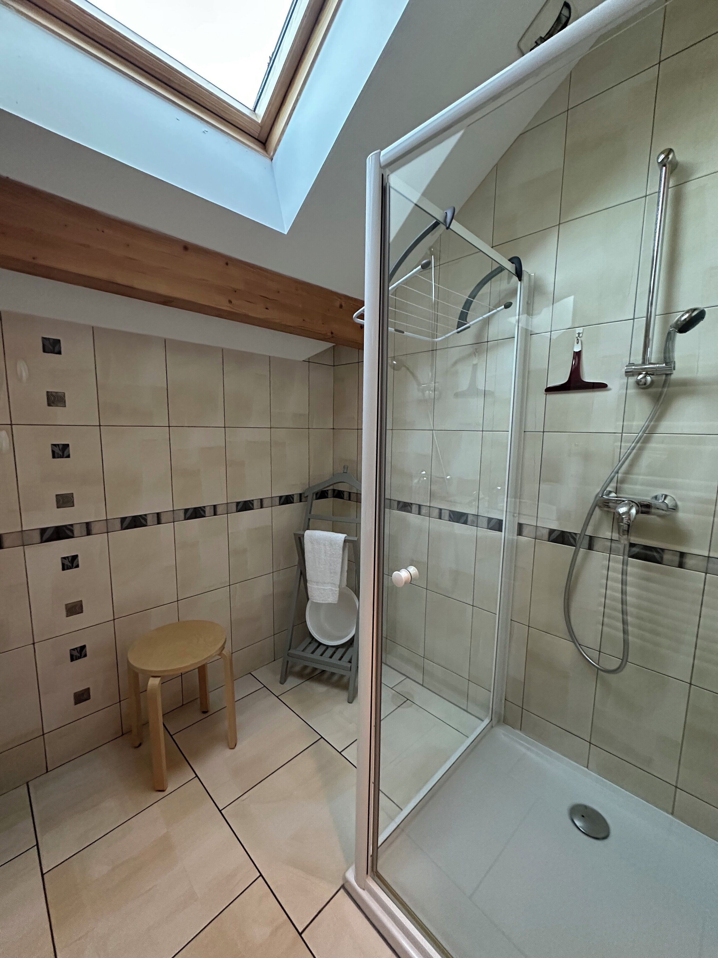 A modern bathroom with a skylight, a glass shower enclosure, a wooden stool, and a towel rack.