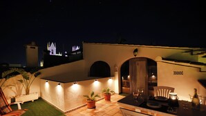 Ein Terrassenbereich im Freien mit Esstisch, Stühlen, Topfpflanzen und Blick auf einen Kirchturm bei Nacht.