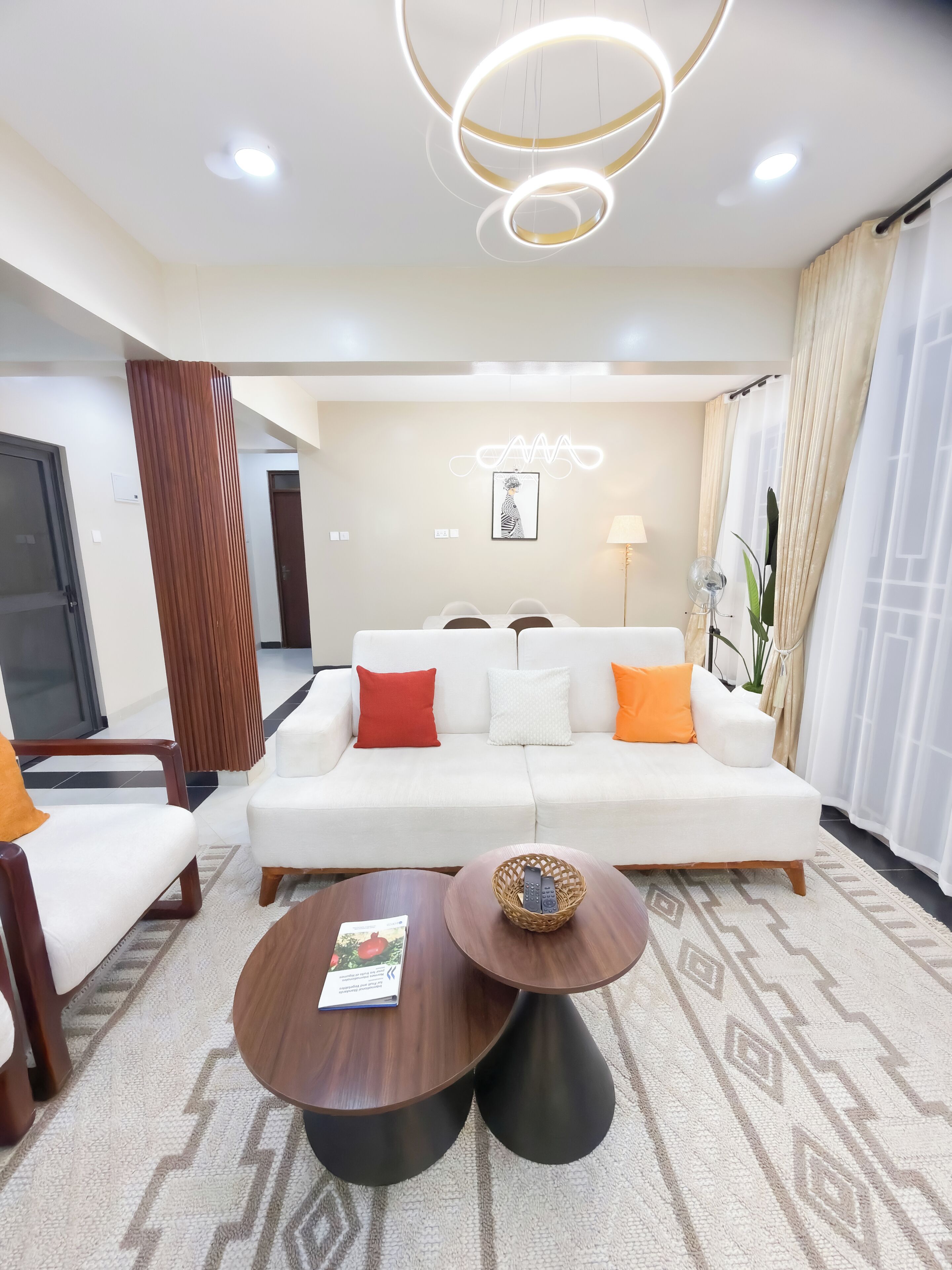 Una sala de estar moderna con un sofá blanco, cojines rojos y blancos, una mesa de centro de madera y una lámpara fijada a la pared.
