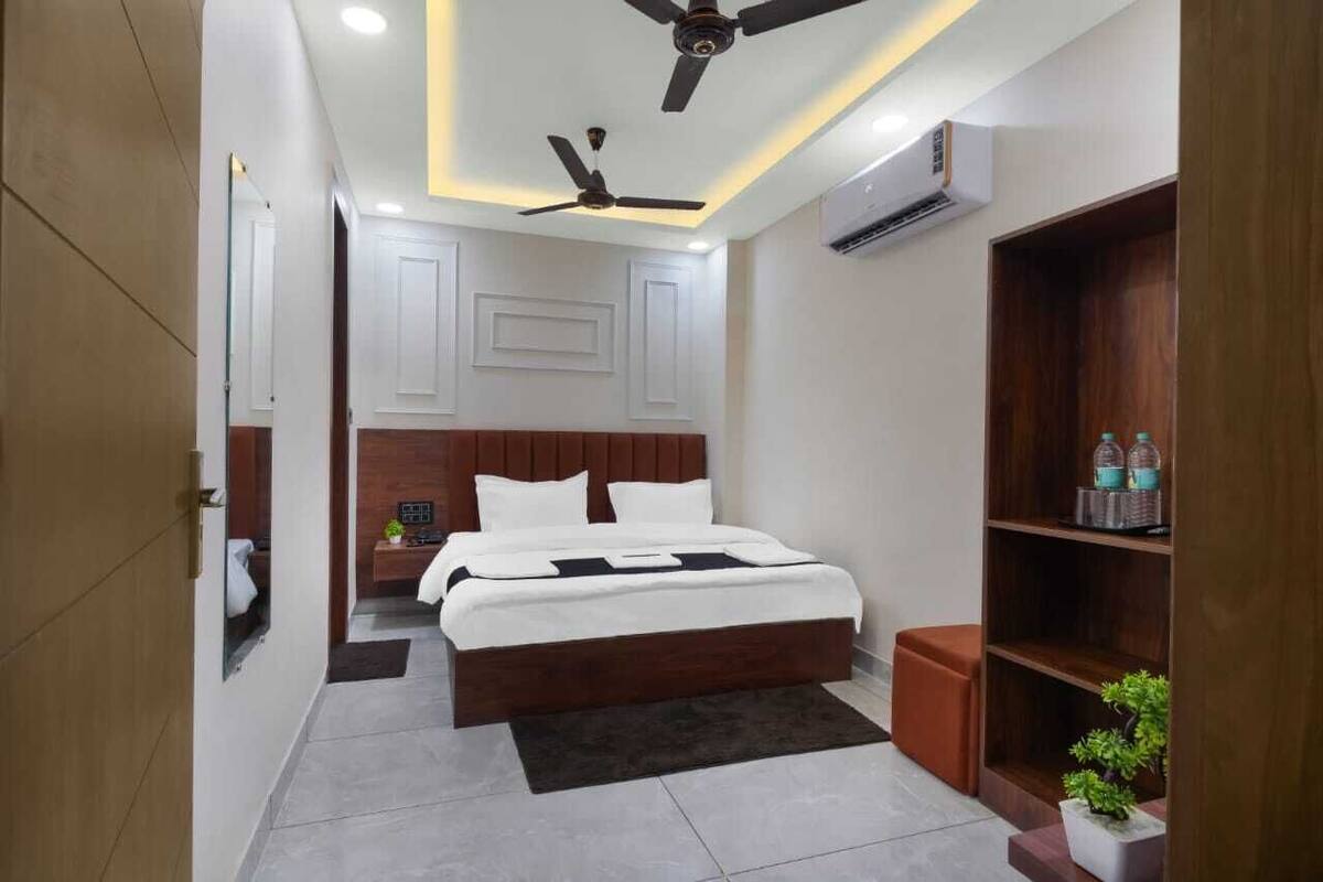 A modern hotel room with a large bed, a ceiling fan, an air conditioning unit, and a wooden bookshelf.