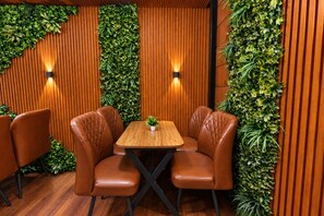 A restaurant interior with a wooden wall, green foliage, leather chairs, and a wooden table. - SG Royal Portico (Haridwar)