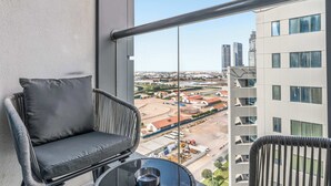 Un balcón con una silla de mimbre y una mesita, con vistas a un paisaje urbano que incluye edificios y una obra en construcción.