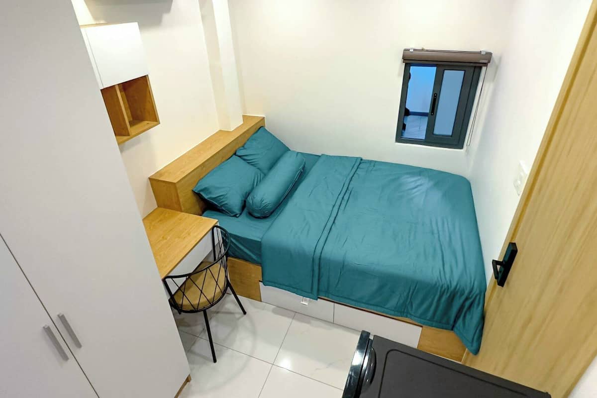 A compact room with a bed, a small desk, a chair, a window, and a door.