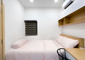 Un dormitorio pequeño y ordenado, con cama, escritorio y ventana con persianas.