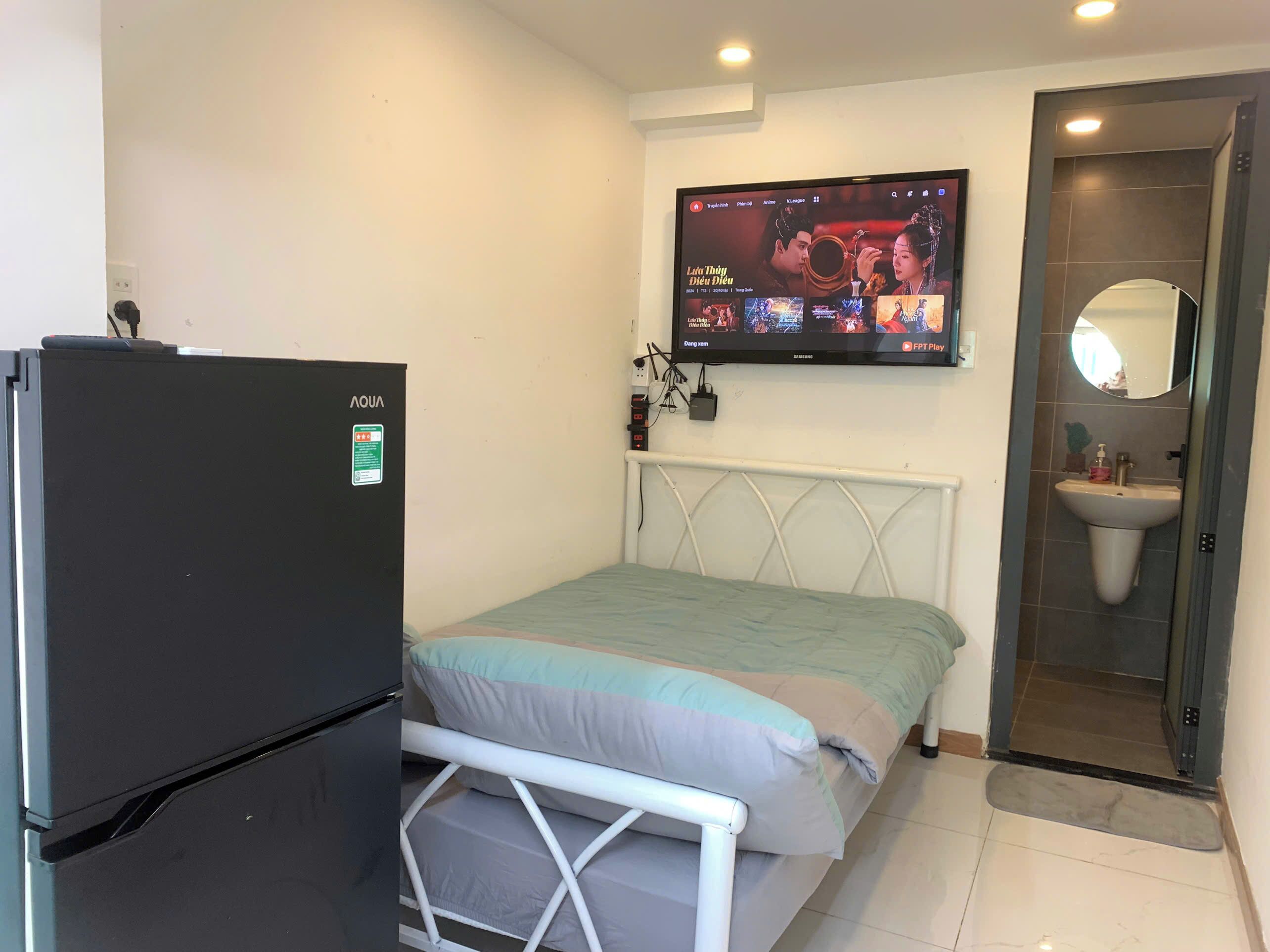 A bedroom with a bed, a TV mounted on the wall, a refrigerator, and a bathroom with a sink and mirror.