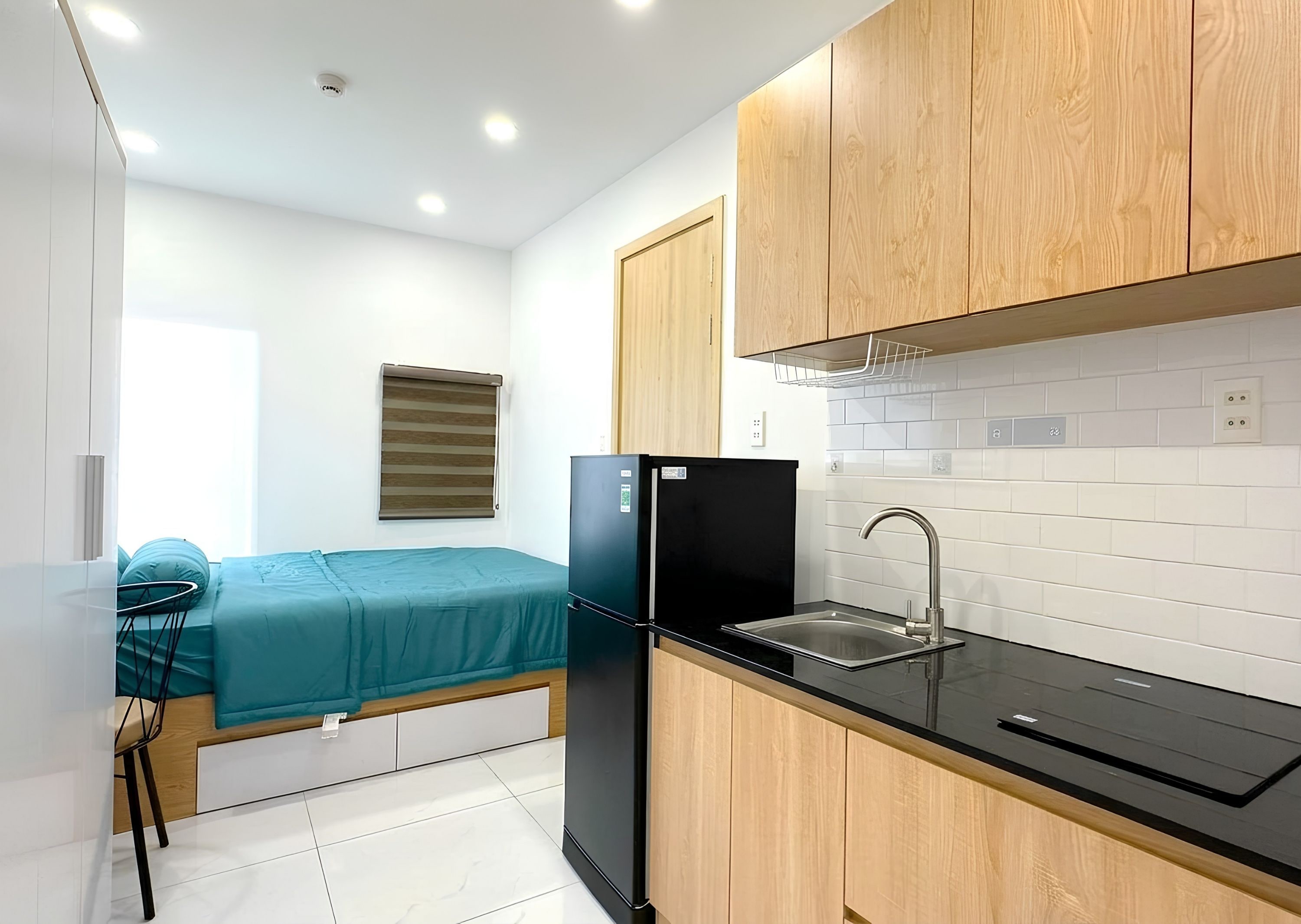 A compact kitchenette with a black refrigerator, wooden cabinets, a sink, and a window with blinds.