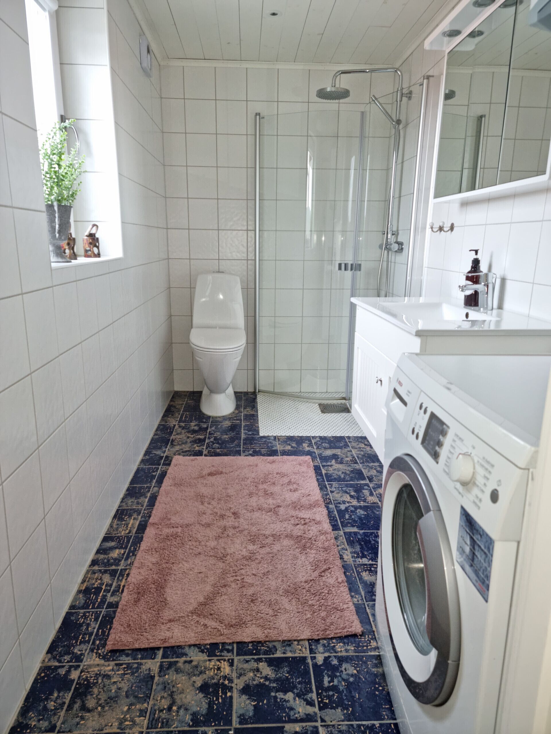 Une petite salle de bain comprenant des toilettes, une douche en verre, un lave-linge et un tapis rose sur le sol.