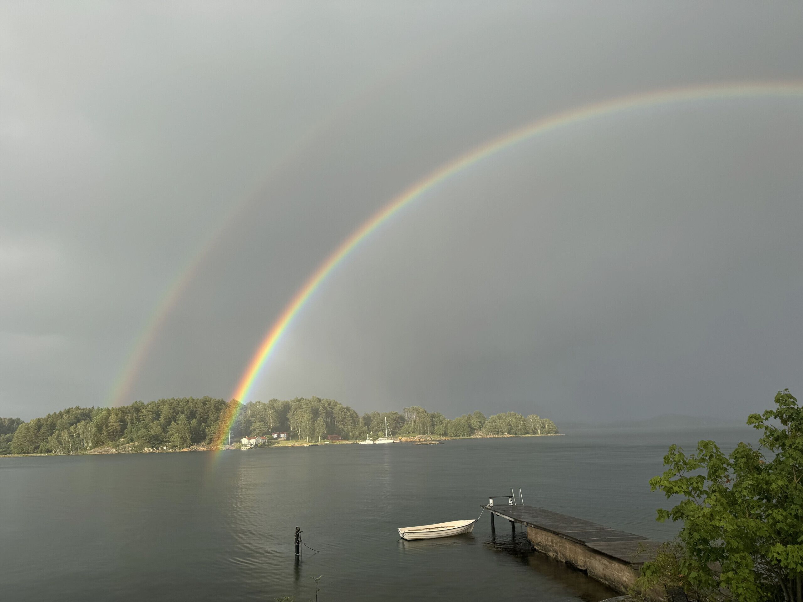 Ein See mit einer Anlegestelle und einem Boot, ein Regenbogen und eine bewaldete Insel im Hintergrund.