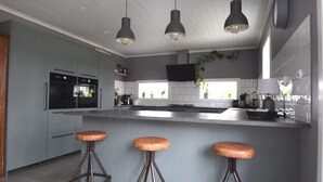 Une cuisine moderne avec des meubles gris, un îlot central doté de tabourets, un éclairage suspendu et un téléviseur à écran plat.