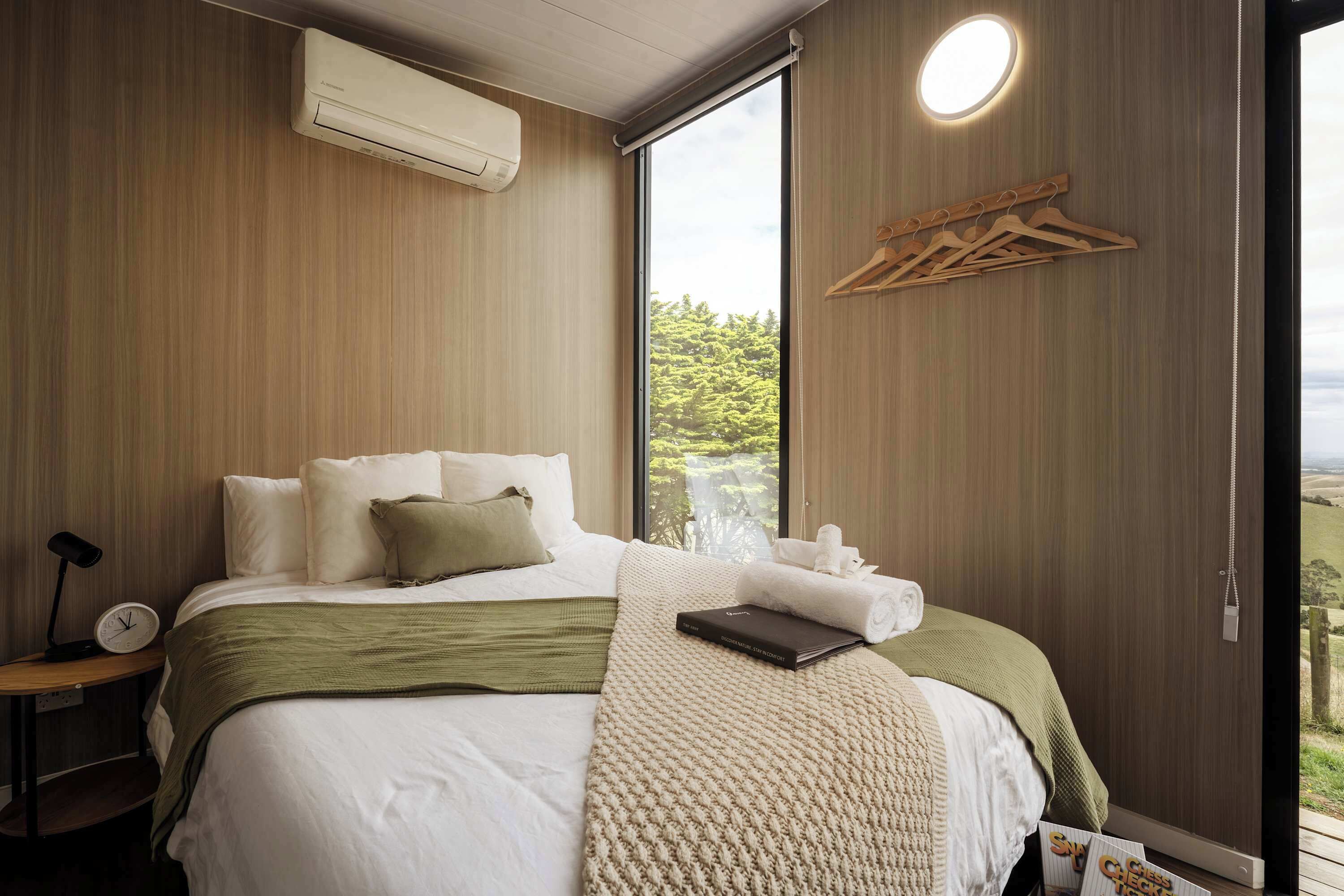 Een slaapkamer met een groot bed, een nachtkastje met een lamp, een wandairconditioner en uitzicht op groen door een groot raam.